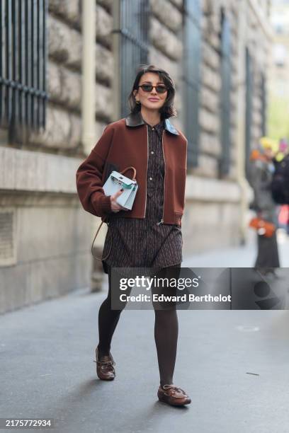 Alexandra Pereira wears silver earrings, dark brown black collar bomber jacket, black pattern mini dress, dark gray sheer mesh tights, shiny dark...