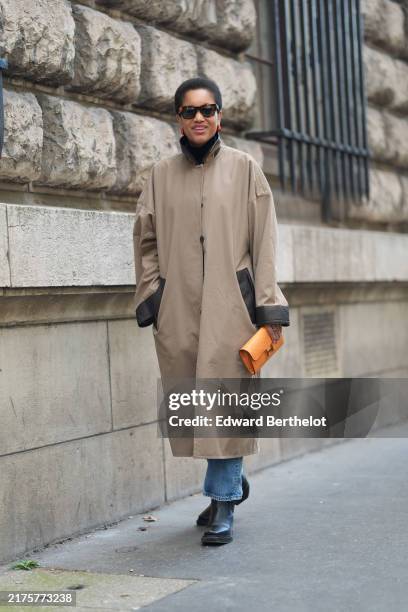 Tamu McPherson wears black sunglasses, light brown oversized trench coat, light blue denim jean pants, shiny black heeled booties leather shoes,...