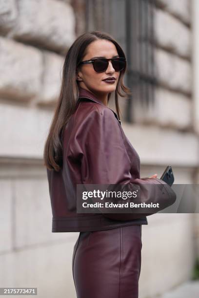 Gabrielle Caunesil wears black sunglasses, shiny dark red leather biker jacket, dark red t-shirt, shiny dark red high waisted high slit leather midi...