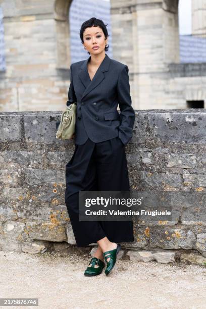 Guest wears gold hoop earrings, a light green leather pouch bag, green leather shoes, a black midi skirt and a grey blazer outside Loewe show during...