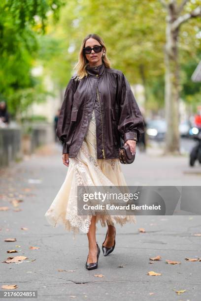 Candela Novembre wears black sunglasses, gold earrings, shiny dark brown oversized bomber leather jacket, cream sheer mesh mermaid flared midi skirt,...