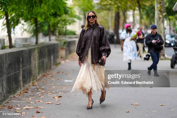 Candela Novembre wears black sunglasses, gold earrings, shiny dark brown oversized bomber leather jacket, cream sheer mesh mermaid flared midi skirt,...