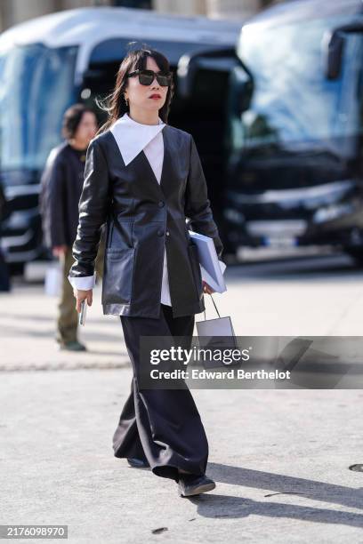 Guest wears black sunglasses, silver earrings, white oversized collar buttoned up long sleeve shirt, shiny black fitted leather blazer jacket, black...