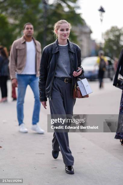 Model wears dark gray sweater, white t-shirt, shiny black oversized leather jacket, black leather belt, dark gray loose pants, shiny black loafer...