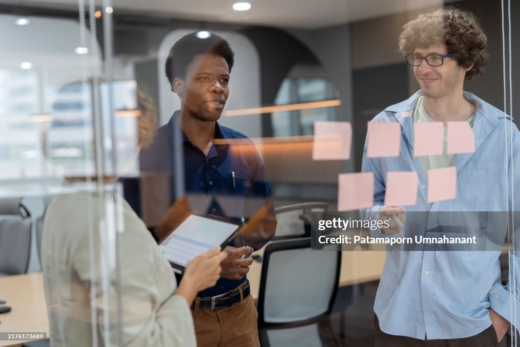 Head of product development standing in a meeting room, discussing ideas with a multiracial team while posting adhesive notes on the glass wall. The session focuses on analyzing market data, creating strategies, and driving business innovation.