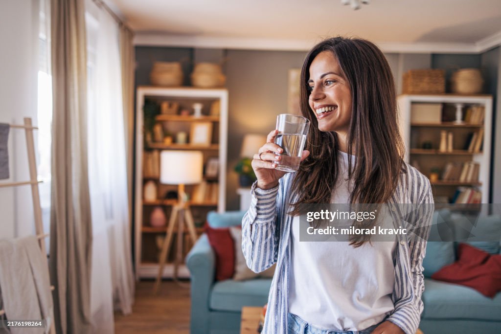 An young cheerful woman standing in the living room and holding glass of water