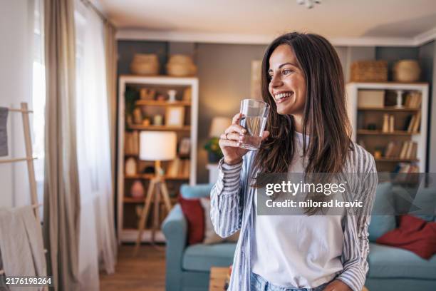 una mujer joven y alegre de pie en la sala de estar y sosteniendo un vaso de agua - agua potable fotografías e imágenes de stock