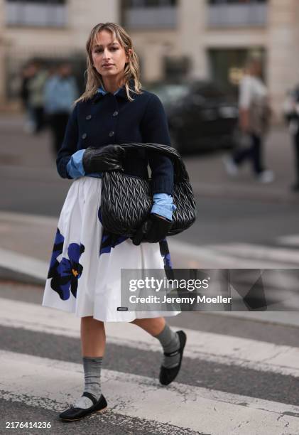 Izzi Allain is seen wearing a cropped dark blue knit cardigan with a double-button placket, a mid blue shirt underneath, a knee-length flared pleated...