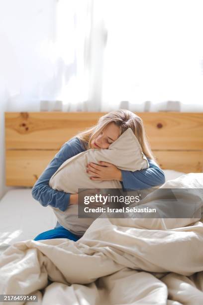 young woman sitting on bed hugging pillow - pijama fotografías e imágenes de stock