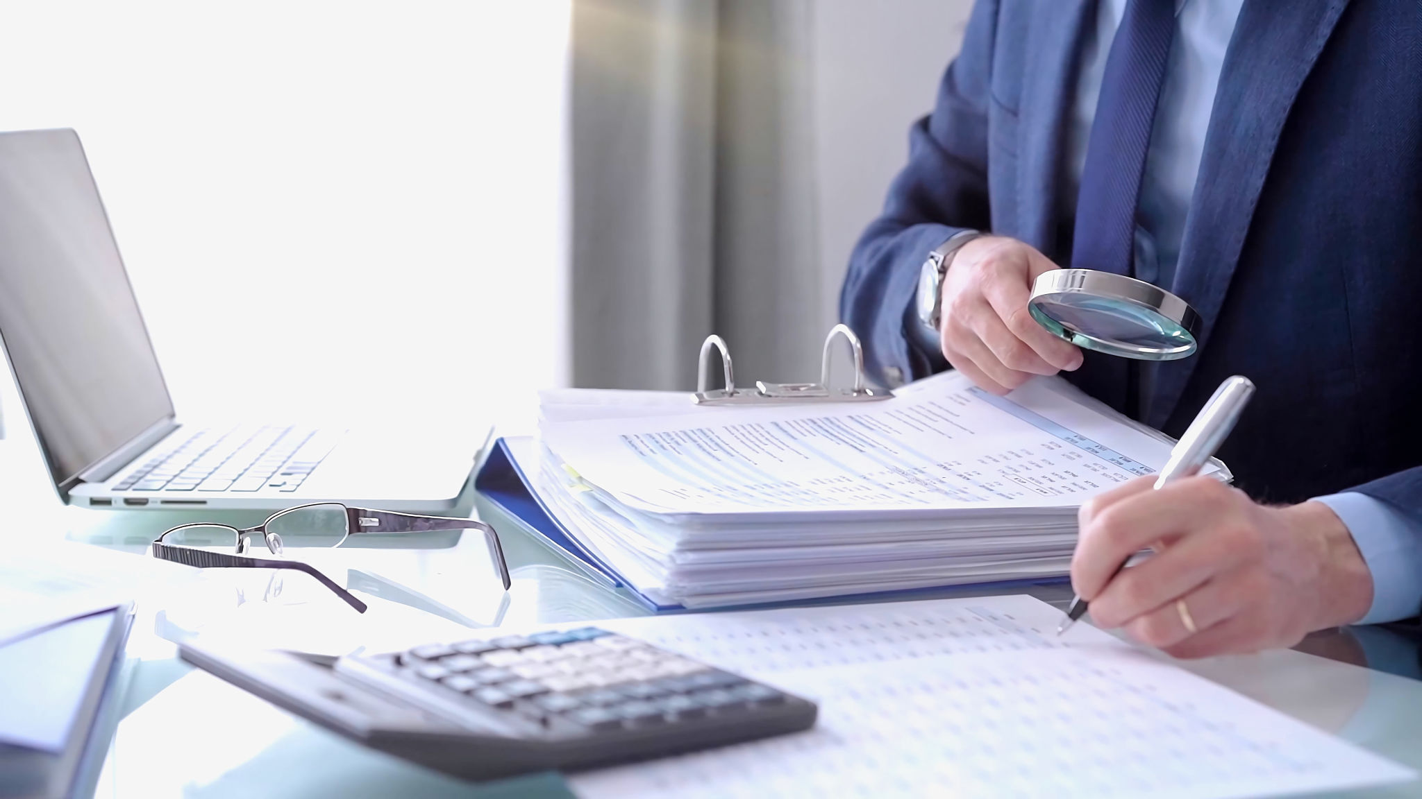Businessman using a calculator and magnifying glass while analyzing financial documents at desk in fair modern office. Audit and taxes in business Businessman using a calculator and magnifying glass while analyzing financial documents at desk in fair modern office. Audit and taxes in business
