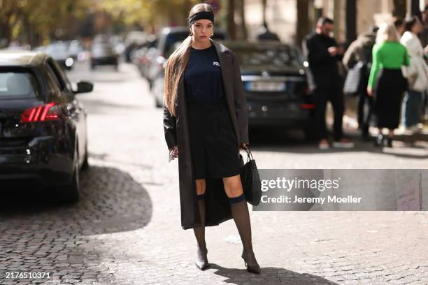 Fashion Week guest is seen wearing a black headband, large gold drop-shaped earrings, a calf-length dark brown leather coat, underneath a dark blue...