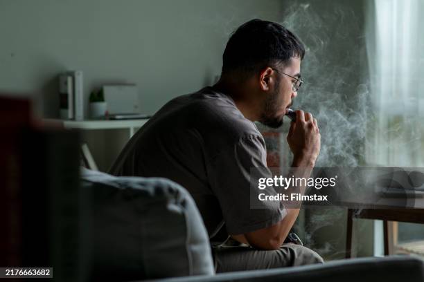 young asian man sitting on a sofa at home, vaping, looking sad and exhausted, struggling with depression and mental health issues. - asian man smoking stock pictures, royalty-free photos & images