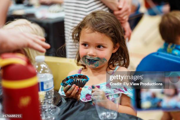 fröhliches mädchen im vorschulalter, das einen cupcake mit blauem zuckerguss im gesicht isst - kuchenboden stock-fotos und bilder