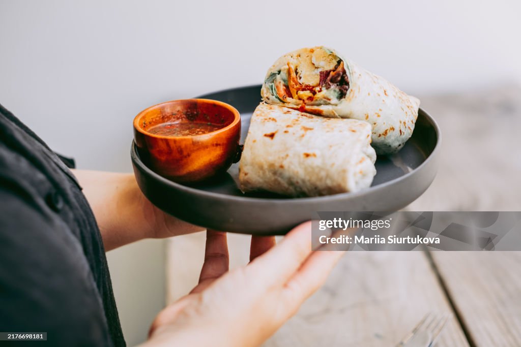 Woman holding plate with vegan burrito with salsa on a rustic wooden table. Alternative eating.