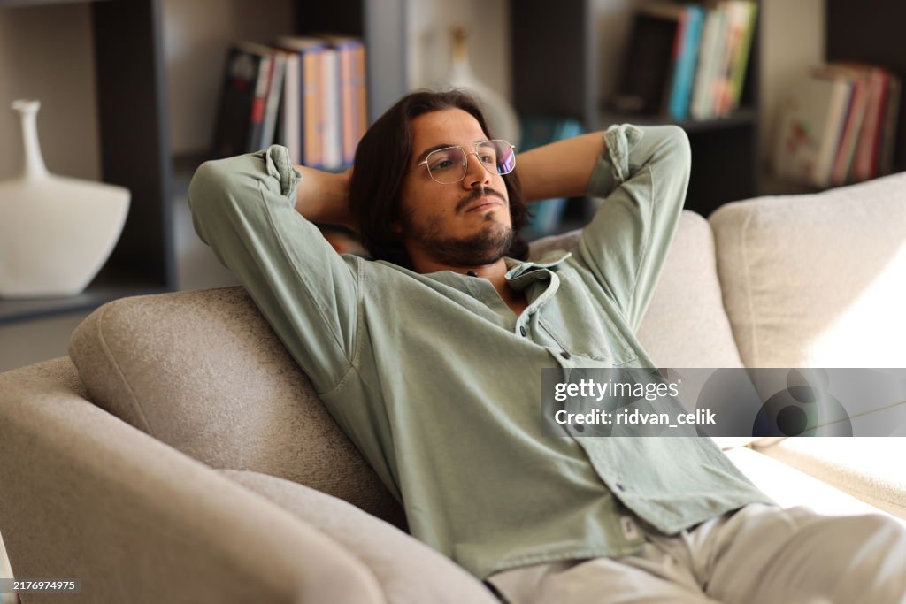 Pensive young man in glasses sitting on sofa at home