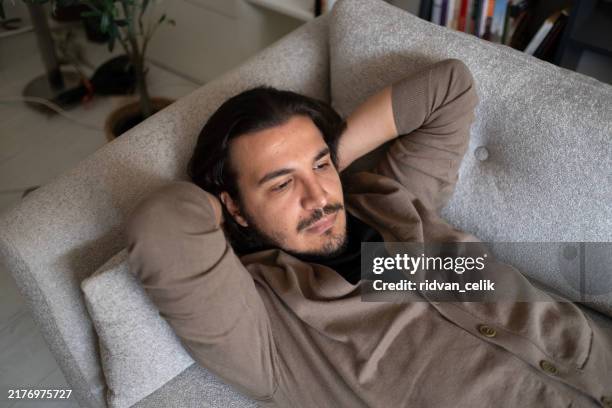pensive young man lying on sofa at home - young widower stock pictures, royalty-free photos & images
