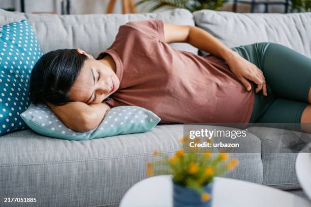 mid-adult pregnant asian woman having contractions while lying on the sofa in the living room - brechreiz stock-fotos und bilder