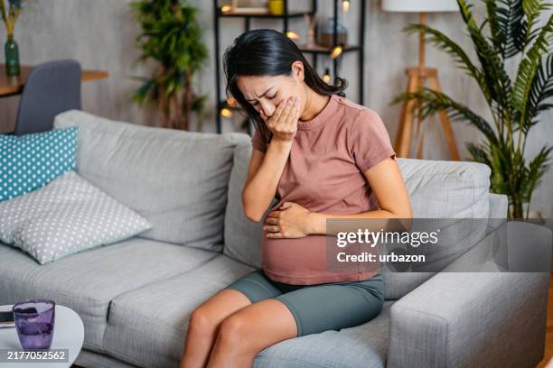 mid-adult pregnant asian woman having morning sickness while sitting on the sofa - muskelkontraktion stock-fotos und bilder