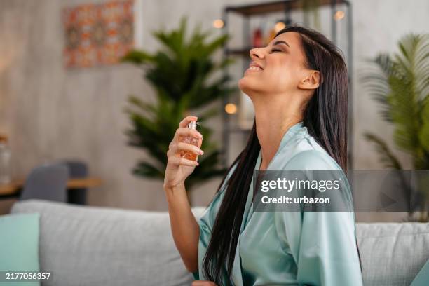 young woman in a bathrobe putting on perfume in the living room - perfumado imagens e fotografias de stock