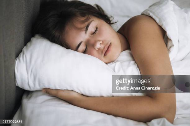 a young person peacefully sleeping in bed under soft white blankets in a cozy room during the morning light - hibernation stock pictures, royalty-free photos & images