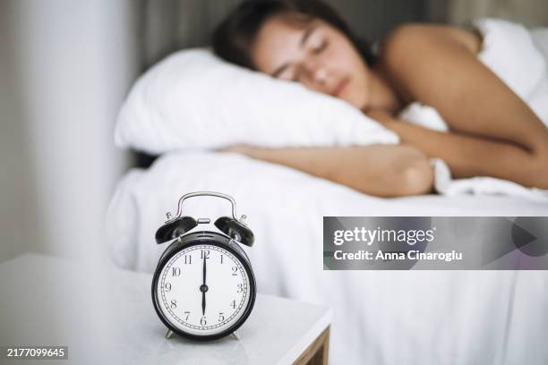 a woman peacefully sleeping in her bedroom, with an alarm clock showing 7 am on a bedside table - hibernation stock pictures, royalty-free photos & images