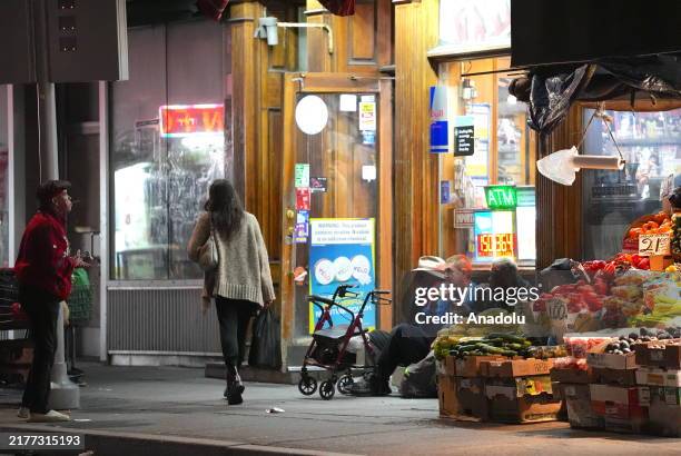 People sit and a woman walks in front of a grocery store in the evening in Greenpoint, Brooklyn in New York City, United States on October 11, 2024.