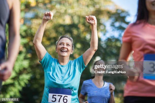 femme mûre levant les mains en célébration du succès tout en courant dans la course de marathon avec un groupe de femmes dans le parc - joggeuse photos et images de collection