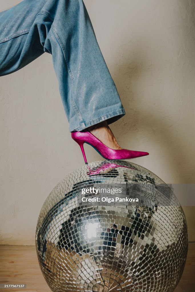 Stylish pink high heel with denim trousers rests on a sparkling disco ball. Playful, nostalgic atmosphere.