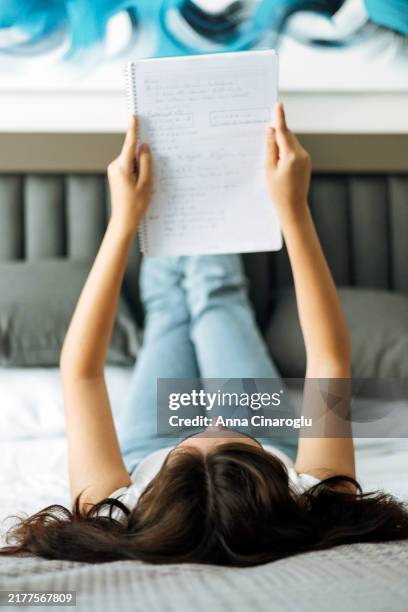 young woman relaxing on a bed while reading a notebook filled with notes in a cozy bedroom setting. student studying on the bed - studentenwohnung stock-fotos und bilder