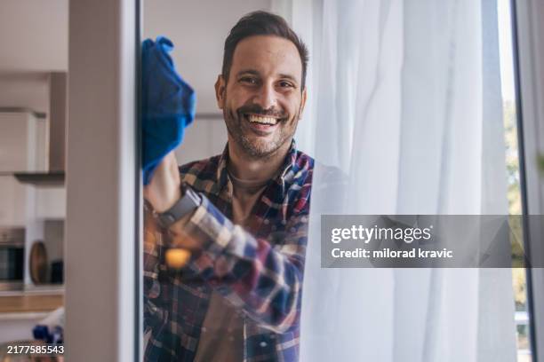 a mid-adult, cheerful, handsome man wiping the windows in the kitchen - cleaning equipment stock pictures, royalty-free photos & images
