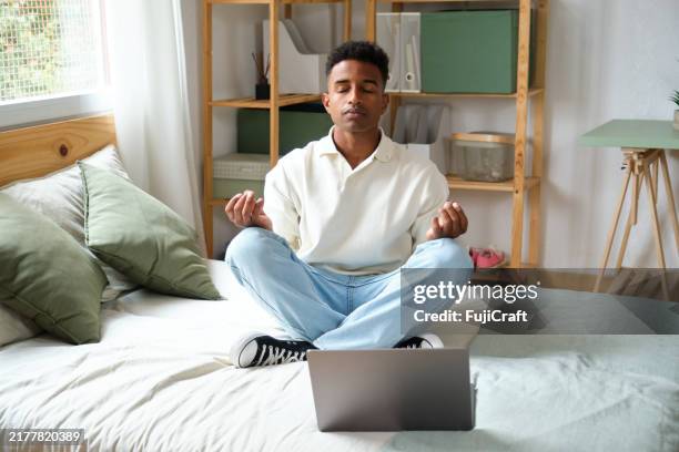 young man meditating in the lotus position on bed with laptop - lotus position stock pictures, royalty-free photos & images