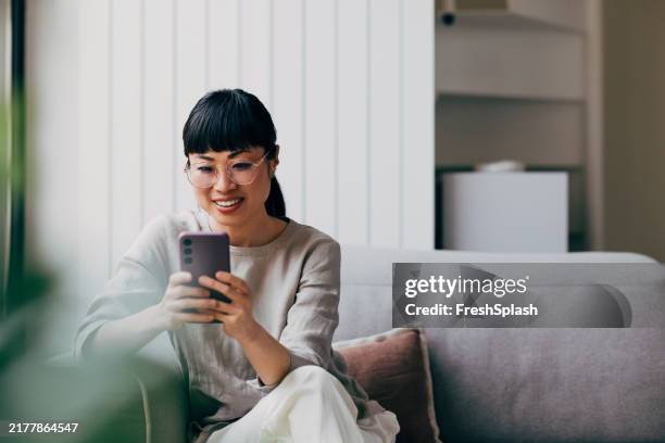 femme heureuse utilisant un smartphone sur un canapé gris moderne à la maison - sourire narquois photos et images de collection