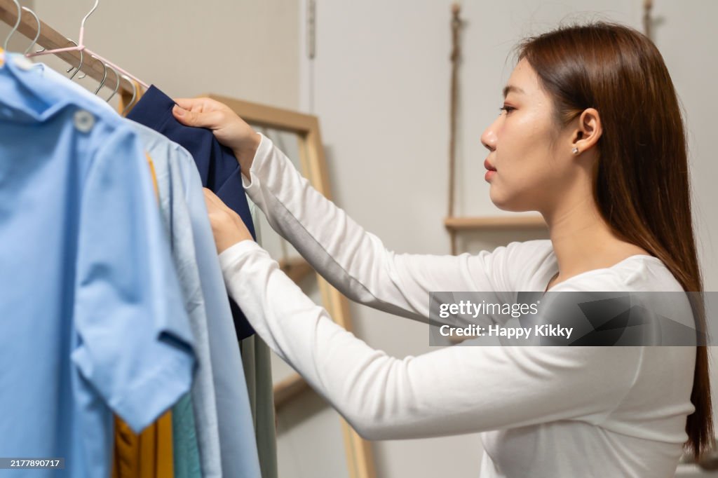 Asian nurse woman takes a hanger with a shirt from clothes rack in the closet bedroom decision-making and personal style in the morning getting ready dressed for work