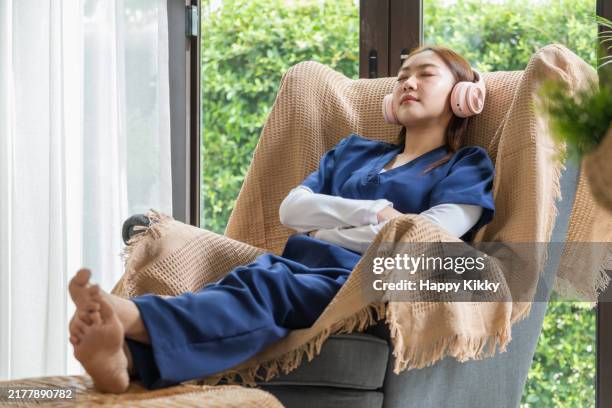 asian woman in blue scrubs uniform wearing pink headphones enjoys a moment of relaxation by listening to music lying on sofa in living room ,taking a break from work - cuidados de saúde primários imagens e fotografias de stock