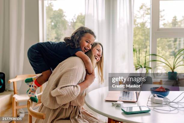 happy mature mother carrying son on shoulders while sitting on chair near table at home - home stockfoto's en -beelden