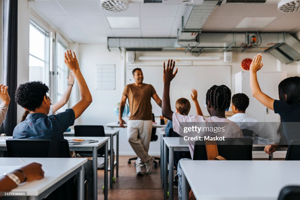 Rear view of male and female junior high students with hand raised while teacher teaching in classroom at school