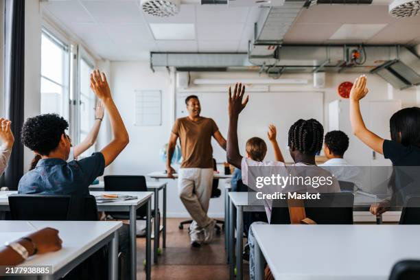 rear view of male and female junior high students with hand raised while teacher teaching in classroom at school - lärare bildbanksfoton och bilder