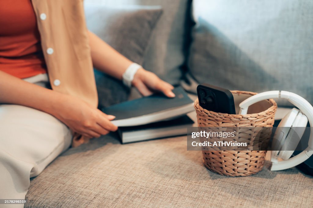 Woman Holding Book Near Basket with Phone and Headphones on Couch