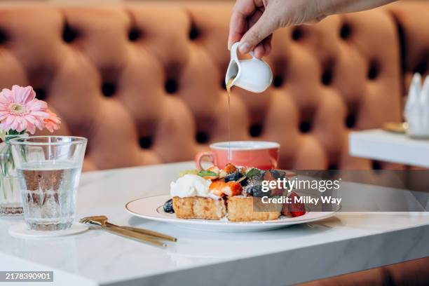 woman pouring syrup over a serving of french toast topped with fresh berries and cream, with a red cup and tufted seating in the background. - toasted bread stock pictures, royalty-free photos & images