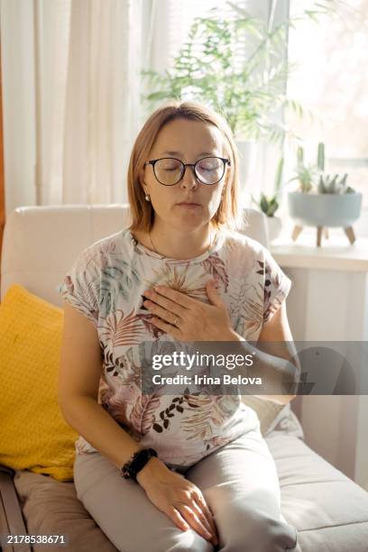 a young woman is sitting on an armchair with her eyes closed, doing breathing exercises, calming herself. - psyche stock pictures, royalty-free photos & images