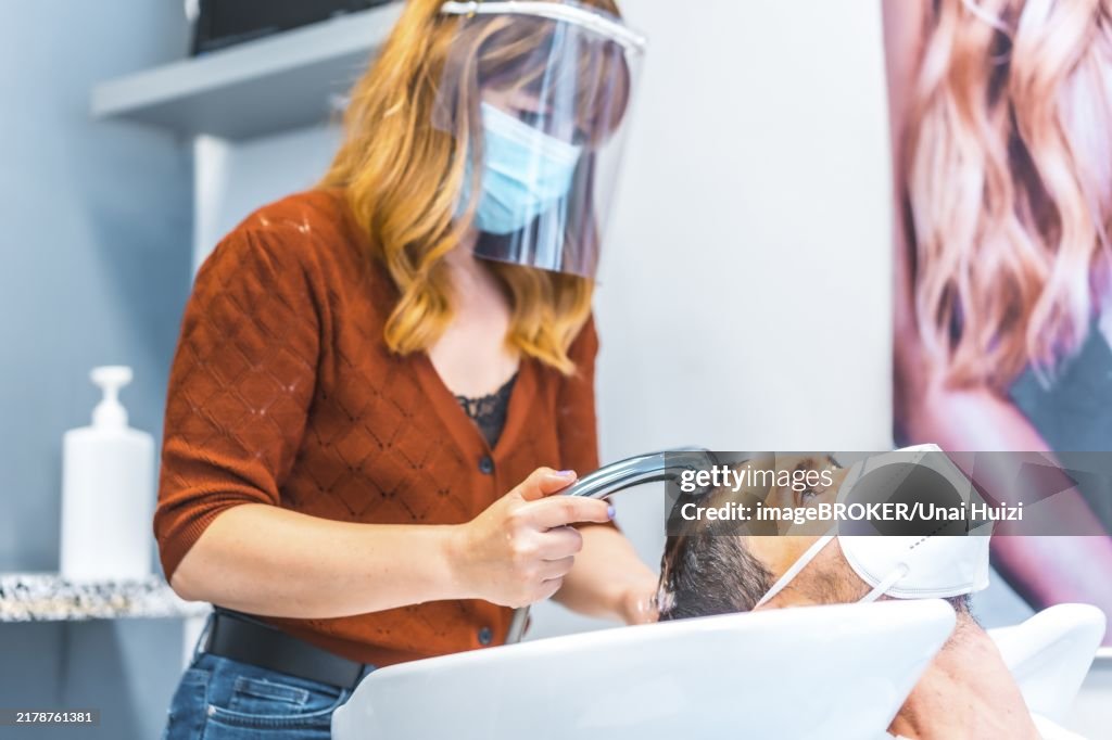 Reopening of hairdressing salons after the Coronavirus pandemic. Hairdresser with face mask and protective screen, covid-19. Social distance, new normality. Washing client's hair with mask