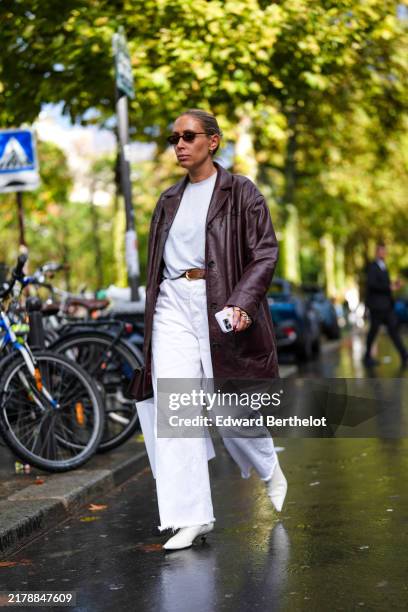 Guest wears black sunglasses, a white top, a dark brown leather coat, a brown leather belt, white pants, white leather pointed shoes high heels,...