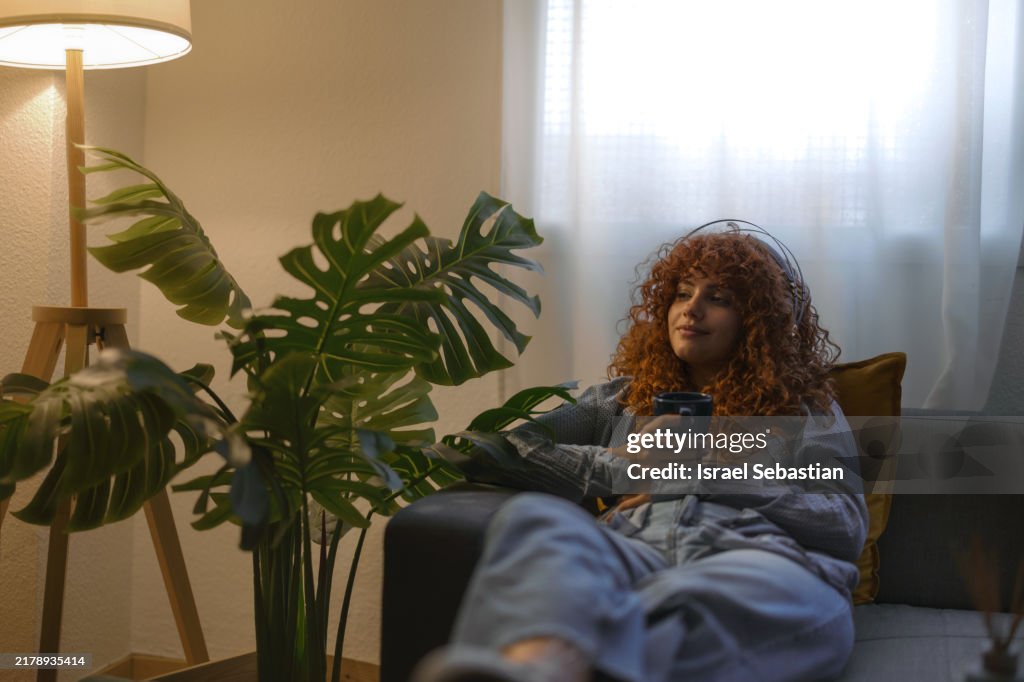 Young woman relaxing on sofa listening music with headphones and drinking coffee