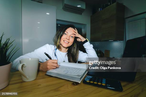 woman writes in a notebook with a laptop, coffee mug and smartphone creating a cozy home office setting for work at night - financial literacy stock pictures, royalty-free photos & images