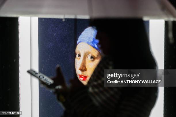 Woman stands in front of a backlit display at a bus stop, with an image of the Girl with a Pearl Earring oil painting by Dutch Golden Age painter...