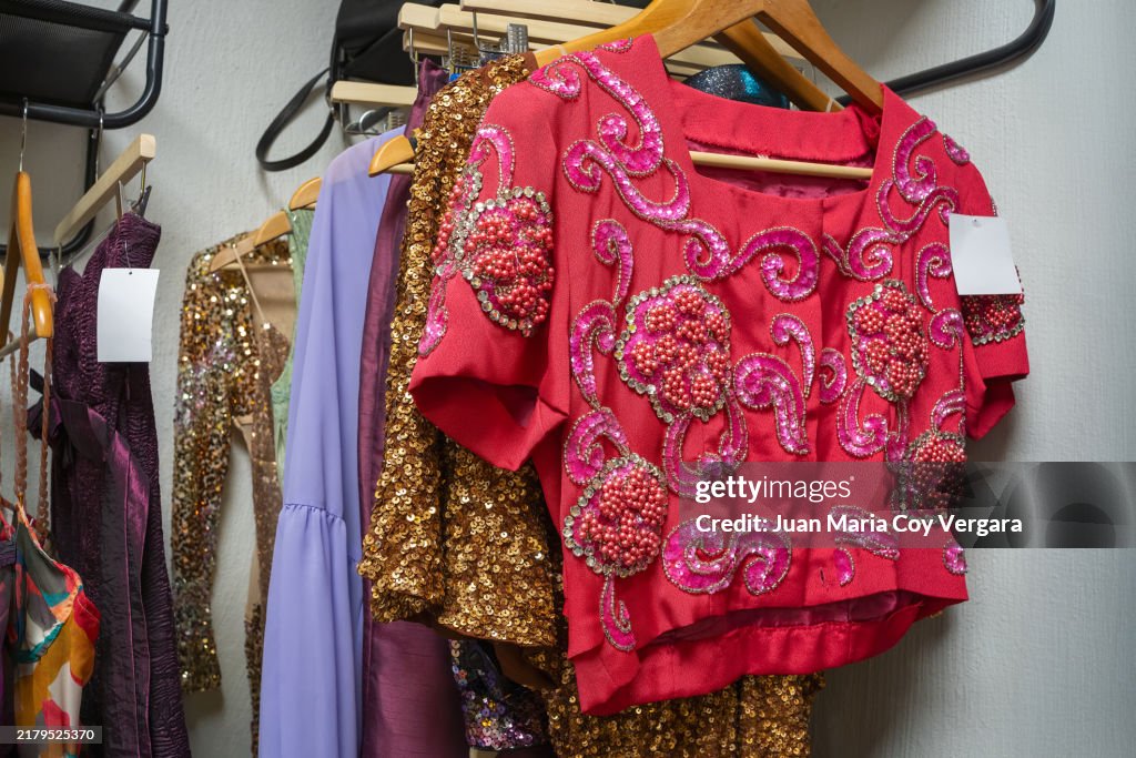A vibrant collection of embellished clothing hangs on wooden hangers in a boutique, centered on a red top embellished with intricate beading and sequins, Spain
