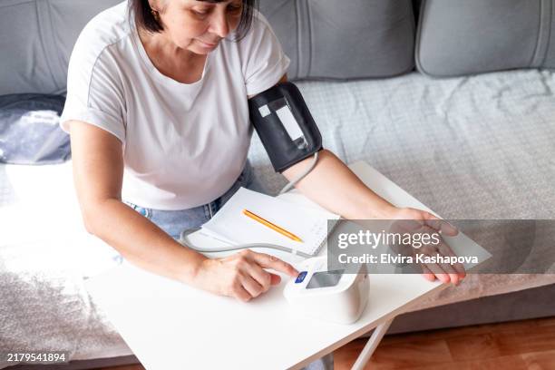 mature woman 58 years old with dark short hair measures blood pressure with a tonometer at a table at home, top view - bluthochdruck stock-fotos und bilder