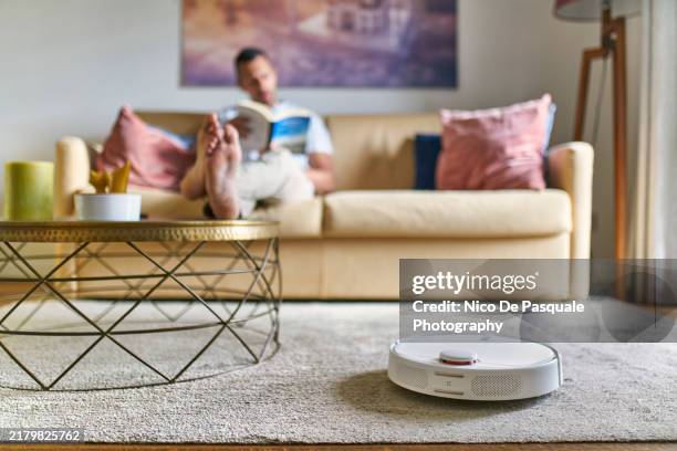 man chilling at home on the sofa while robot vacuum cleaner is working - robot cleaner stock pictures, royalty-free photos & images