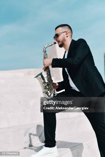 talented male saxophonist showcasing his talent and passion while playing saxophone outdoors dressed in black suit - saxophone stock pictures, royalty-free photos & images