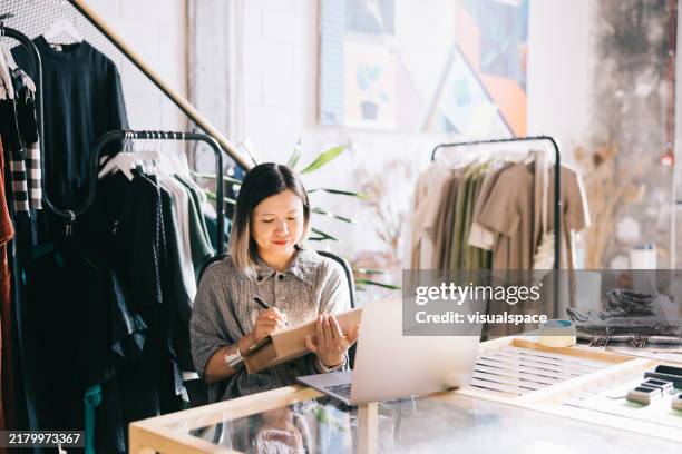 la proprietaria di un negozio di abbigliamento si siede nel suo spazio di lavoro con il suo laptop aperto, scrivendo l'indirizzo del cliente su una scatola - studio di design foto e immagini stock